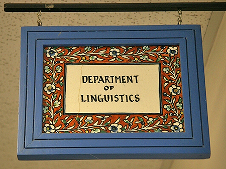 A hanging sign with a blue border and interior tiled decoration with flowers and a rectanagle reading 'Department of Linguistics'